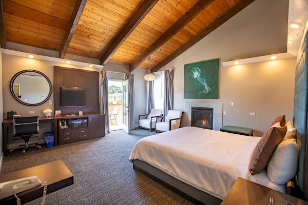 A modern bedroom with a bed, TV, desk, chairs, and a fireplace, featuring wooden ceilings and a large window with curtains.