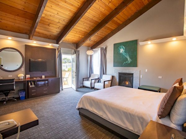 A modern bedroom with a bed, TV, desk, chairs, and a fireplace, featuring wooden ceilings and a large window with curtains.