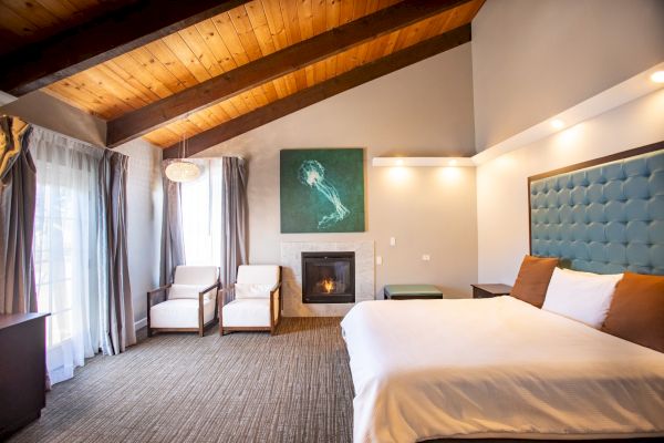 This image shows a cozy bedroom with a sloped wooden ceiling, large bed, chairs, fireplace, and a window with curtains, creating a warm ambiance.