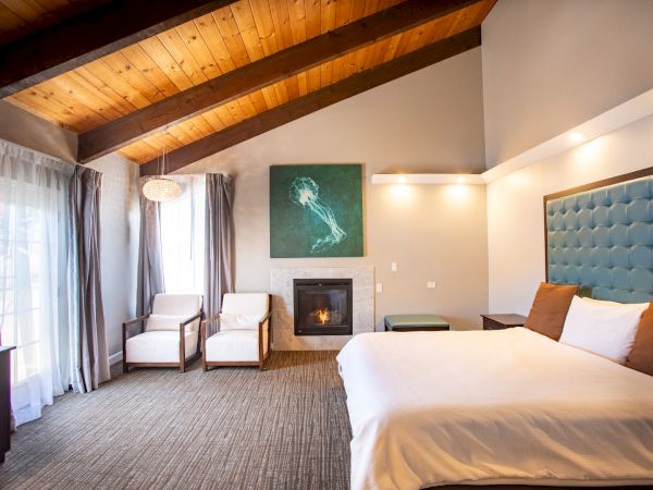 This image shows a cozy bedroom with a sloped wooden ceiling, large bed, chairs, fireplace, and a window with curtains, creating a warm ambiance.