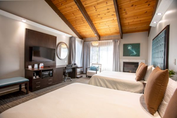 A cozy hotel room with two beds, a TV, desk, and seating area. Features wooden ceiling, fireplace, and large windows.