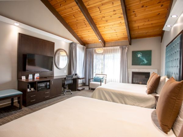 A cozy hotel room with two beds, a TV, desk, and seating area. Features wooden ceiling, fireplace, and large windows.
