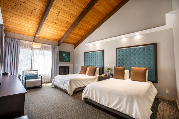 A cozy hotel room with two beds, tufted headboards, wooden ceiling, armchair, and a large window allowing natural light.