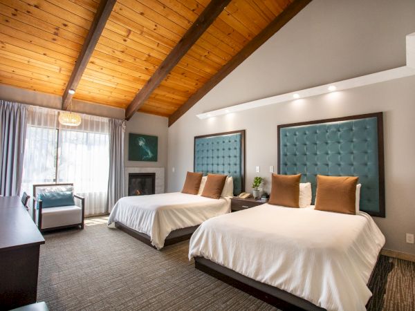 A cozy hotel room with two beds, tufted headboards, wooden ceiling, armchair, and a large window allowing natural light.