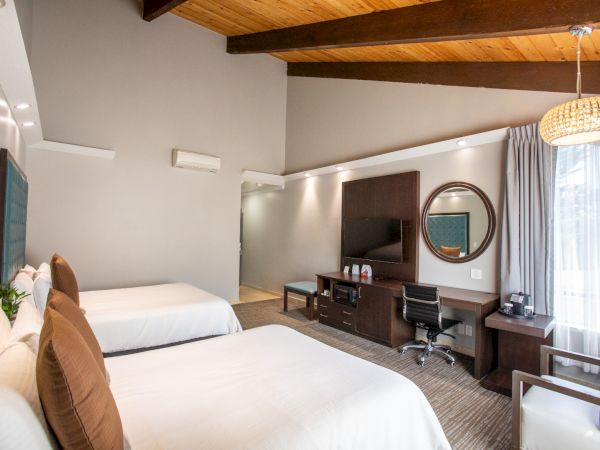 A hotel room with two beds, a desk, TV, seating area, and large windows. It features modern decor and wooden ceiling beams.