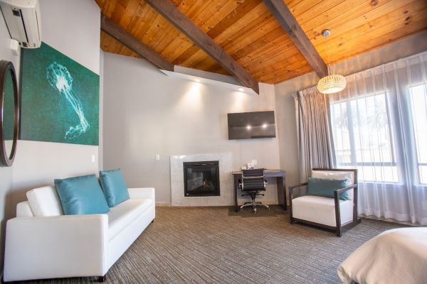 A cozy room with a white sofa, teal pillows, desk, chair, wall art, TV, fireplace, large window, and wood ceiling.