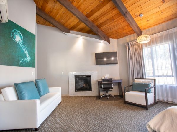 A cozy room with a white sofa, teal pillows, desk, chair, wall art, TV, fireplace, large window, and wood ceiling.