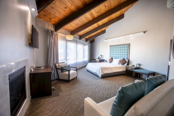 A spacious hotel room features a bed, sofa, fireplace, desk, and TV, with wooden ceiling beams and large windows enhancing the ambiance.