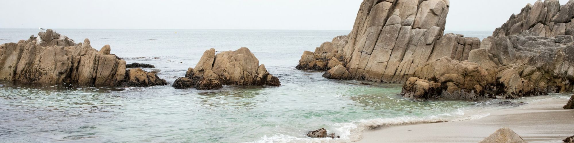 A sandy beach with gentle waves, rocks in the foreground and background, and a calm sea under a cloudy sky.