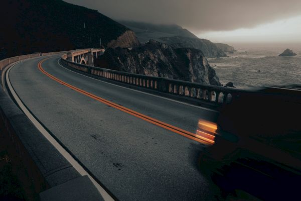 A winding coastal road with light trails from passing vehicles, set against a backdrop of dramatic clouds and rocky cliffs by the ocean.