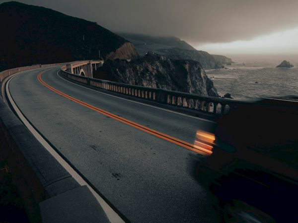 A winding coastal road with light trails from passing vehicles, set against a backdrop of dramatic clouds and rocky cliffs by the ocean.