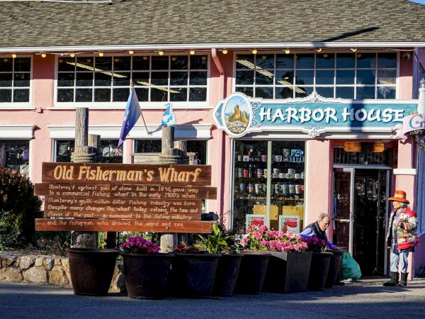 A pink building with "Harbor House" signage, flags, and a large wooden sign for "Old Fisherman's Wharf" in front, surrounded by planters.