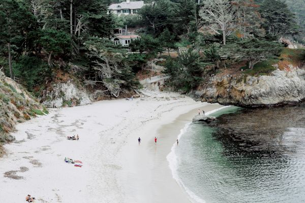 A serene beach scene with a sandy shoreline, few people walking, and forested cliffs in the background. Overcast sky sets a peaceful tone.