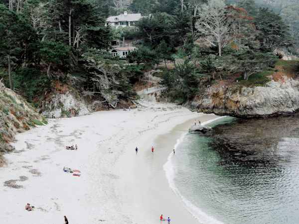 A serene beach scene with a sandy shoreline, few people walking, and forested cliffs in the background. Overcast sky sets a peaceful tone.