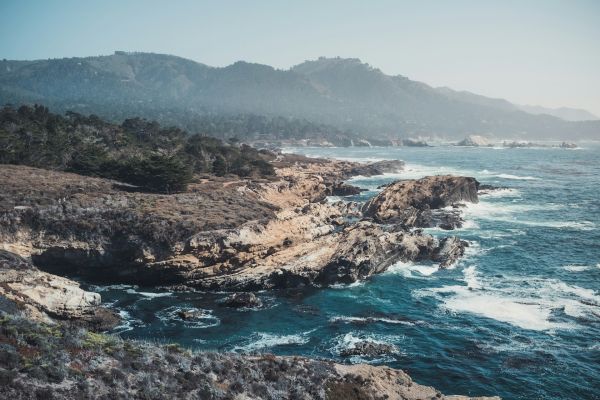 A rocky coastline with waves crashing against the shore, surrounded by rugged cliffs and distant mountains under a clear sky.