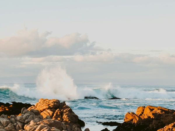 A rocky shoreline with waves crashing against the rocks under a partly cloudy sky during sunset or sunrise.