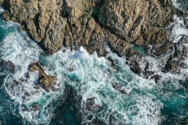 Waves crash against rugged rocks on a coastline, creating splashes and foam in a dynamic seascape captured from above.