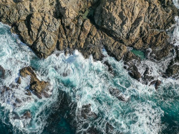 Waves crash against rugged rocks on a coastline, creating splashes and foam in a dynamic seascape captured from above.