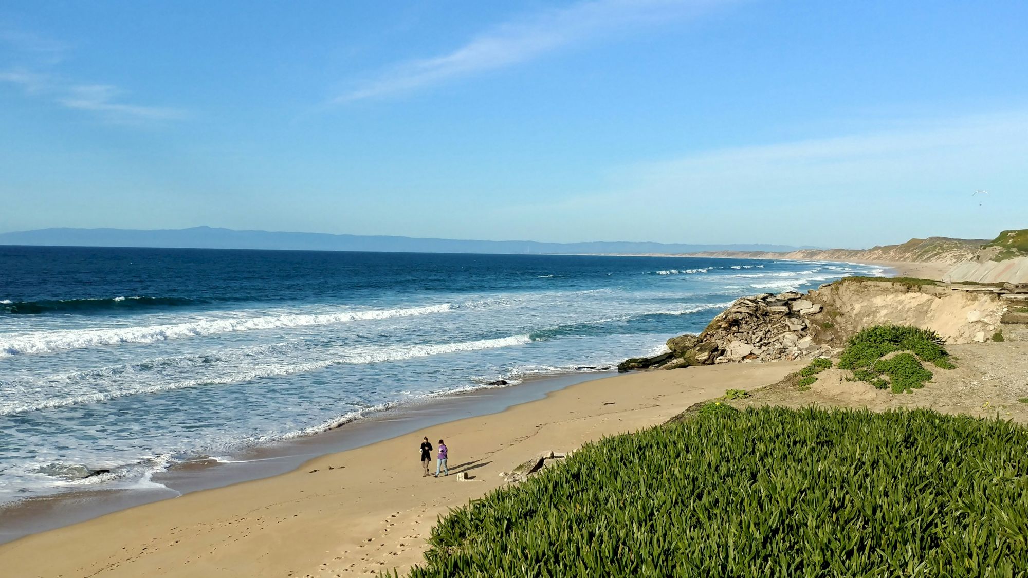 A vast beach with gentle waves, a rocky coastline, and green vegetation. Two people stroll along the sandy shore in the serene setting.