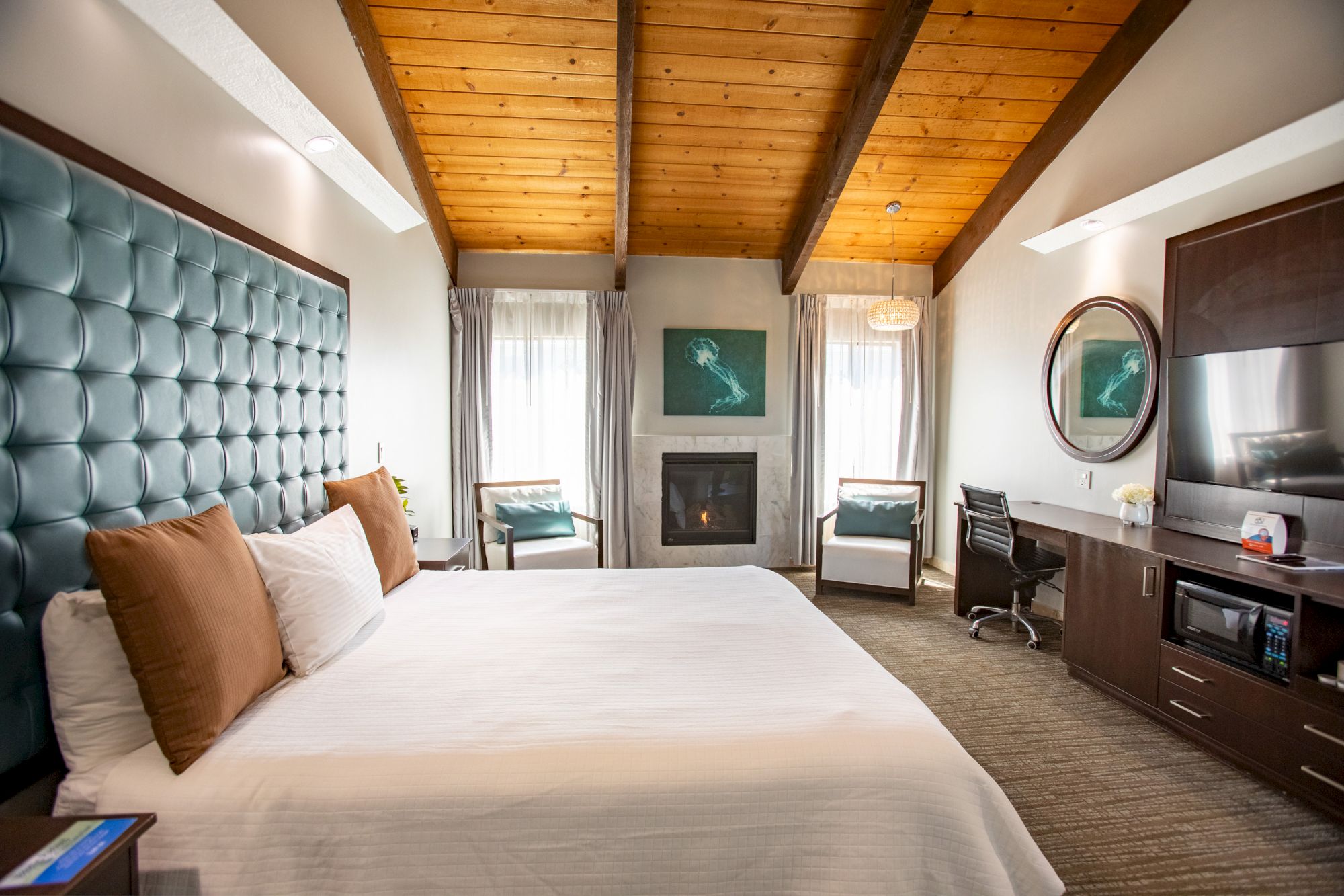 A cozy hotel room with a wooden ceiling, king-size bed, fireplace, TV, desk and chairs, decorated with turquoise and brown accents.