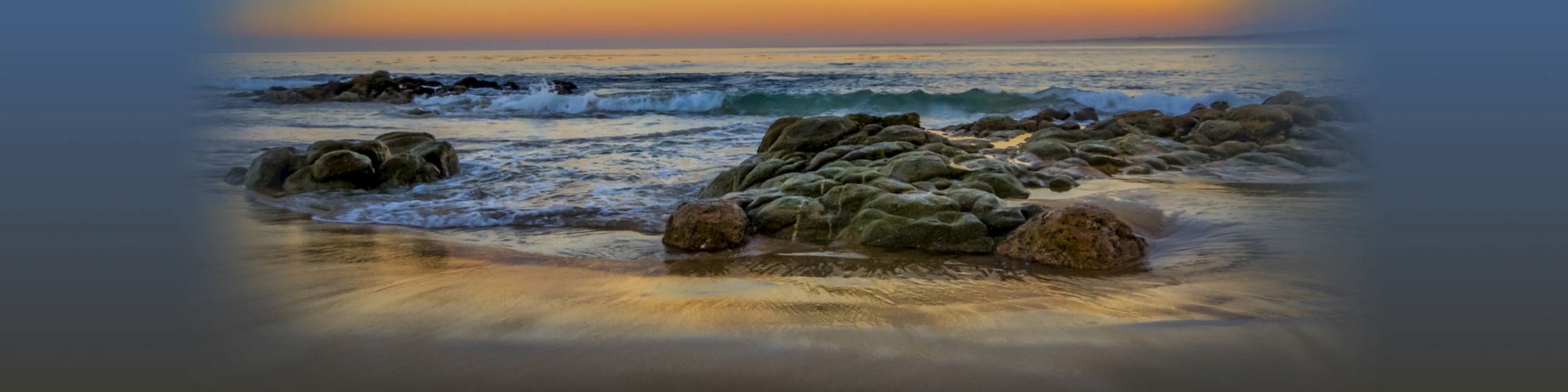 A serene beach scene at sunset with gentle waves lapping against rocky formations and a glowing horizon.