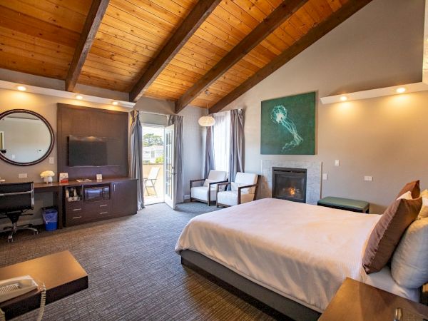 A cozy hotel room features a bed, desk, TV, fireplace, and seating area, with wooden ceilings and natural light from the open door.