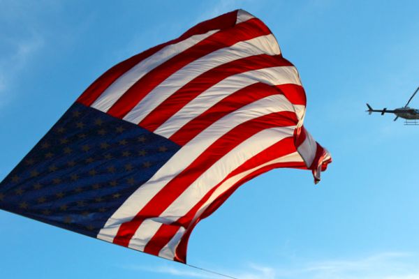 A large American flag waves in the sky alongside a helicopter flying against a blue backdrop.