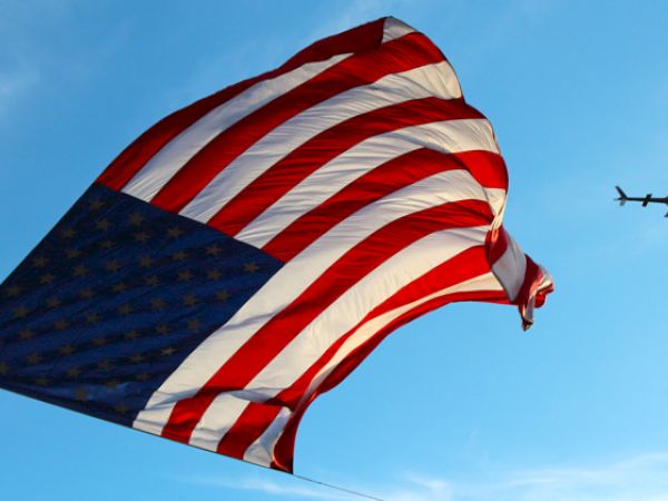 A large American flag waves in the sky alongside a helicopter flying against a blue backdrop.