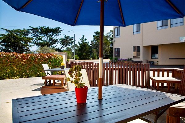 A patio with a wood table, a red potted plant, and a blue umbrella. Fenced seating area near a building with greenery in the background.