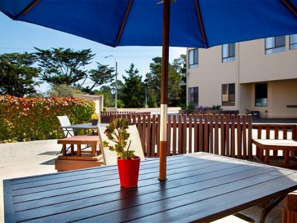 A patio with a wood table, a red potted plant, and a blue umbrella. Fenced seating area near a building with greenery in the background.