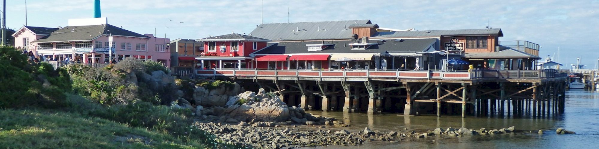 A waterfront scene with buildings on stilts, rocky shoreline, and vibrant purple flowers in the foreground, under a clear blue sky.