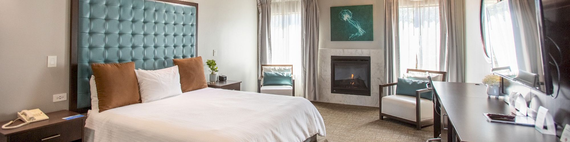 A cozy bedroom with a wooden ceiling, bed with blue headboard, fireplace, armchair, and desk, creating a warm ambiance.