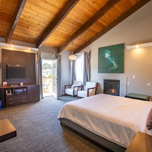 A cozy bedroom with a vaulted wooden ceiling, featuring a bed, desk, fireplace, TV, and a door leading outside, near two chairs.