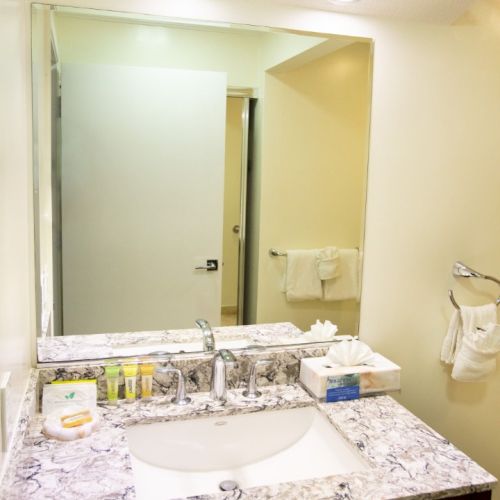 A bathroom vanity with a marble countertop, mirror, sink, and toiletries. Towels hang on the wall to the right.