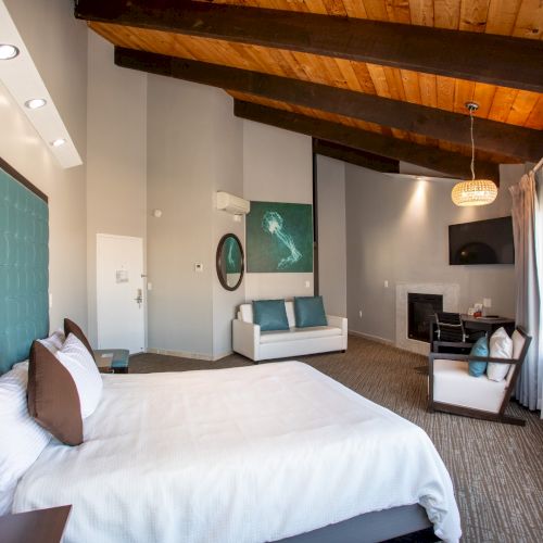 A modern bedroom with a vaulted wooden ceiling, turquoise headboard, white bedding, and a cozy seating area by a fireplace.