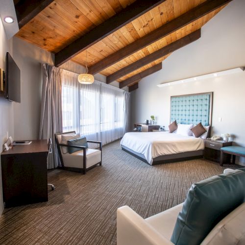 A cozy hotel room with a bed, sofa, fireplace, desk, and large window, featuring exposed wood beams and modern decor.