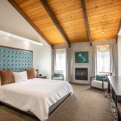 A cozy bedroom with a wooden ceiling, bed, fireplace, chairs, desk, and TV. Neutral tones and modern furnishings create a relaxing ambiance.