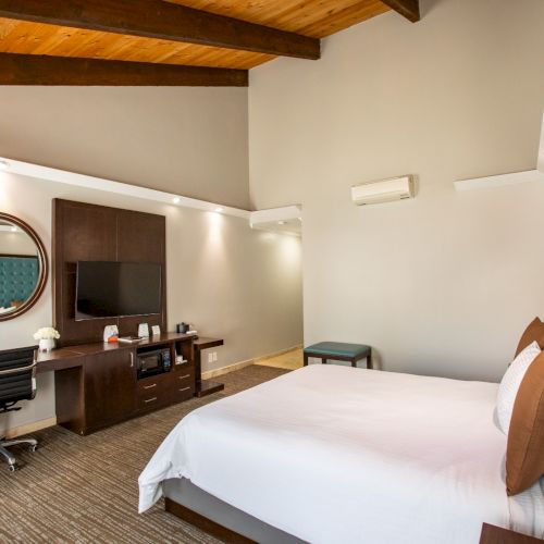 This image shows a modern hotel room with a bed, desk, TV, and decor. The room has wooden beams and a plant near the bed.