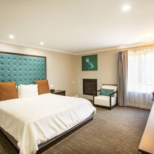 A modern hotel room with a large bed, teal headboard, desk, chair, and window with curtains. Warm lighting and neutral tones create a cozy ambiance.