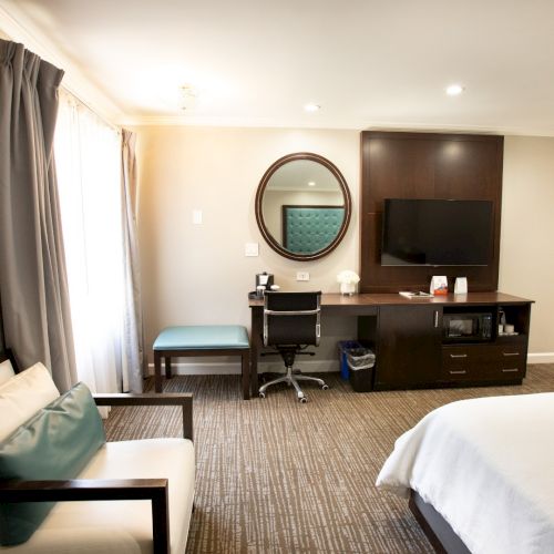 The image shows a hotel room with a bed, a desk, chair, wall-mounted TV, a mirror above the desk, and a small couch by the window.