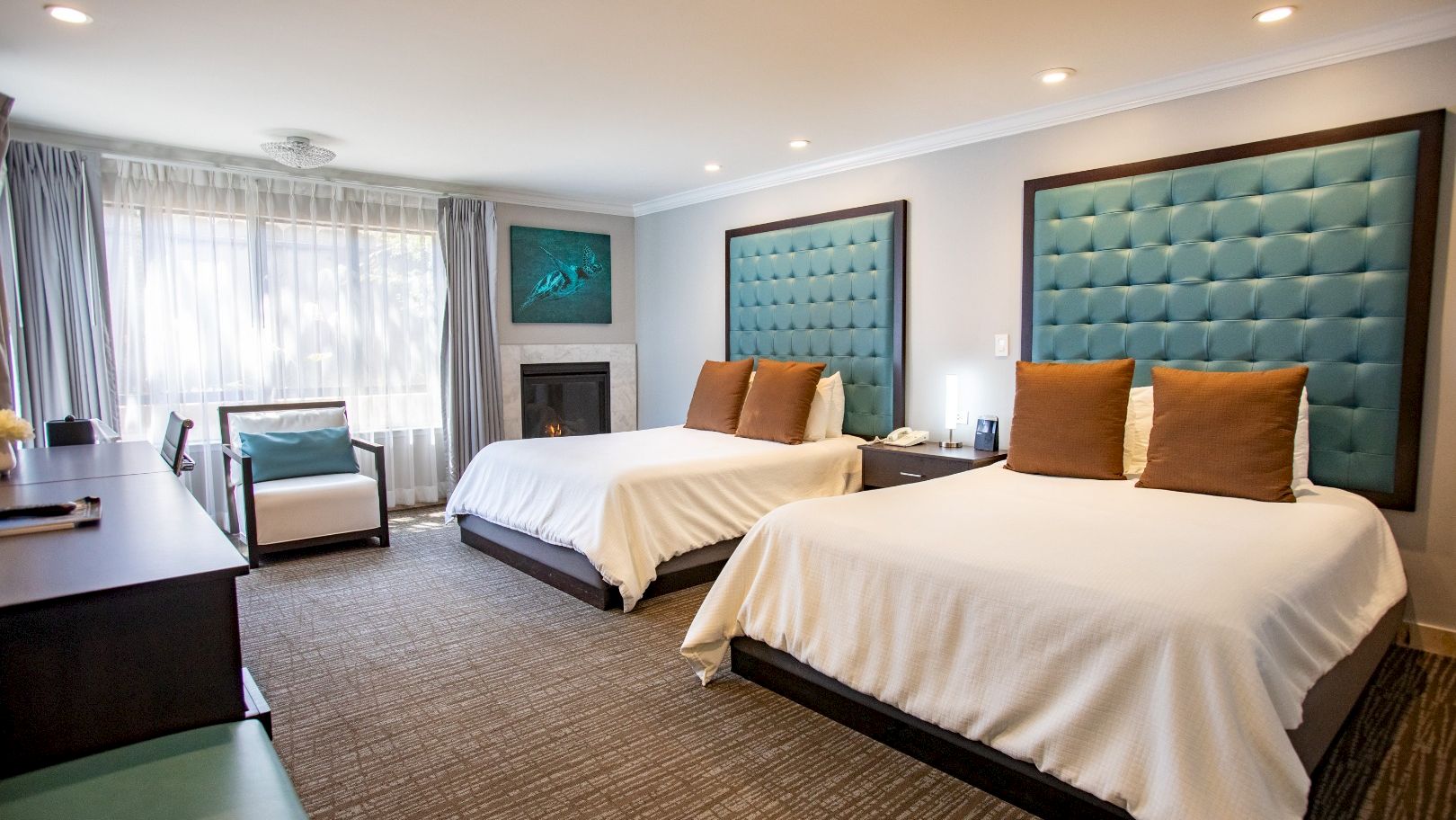 A hotel room with two beds, teal padded headboards, a desk, chair, and large window with curtains. Neutral tones and modern decor style.