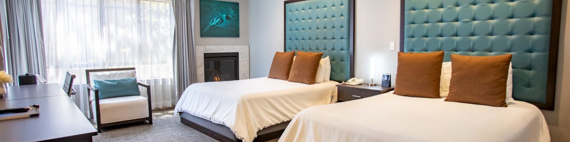 A hotel room with two beds, teal padded headboards, a desk, chair, and large window with curtains. Neutral tones and modern decor style.