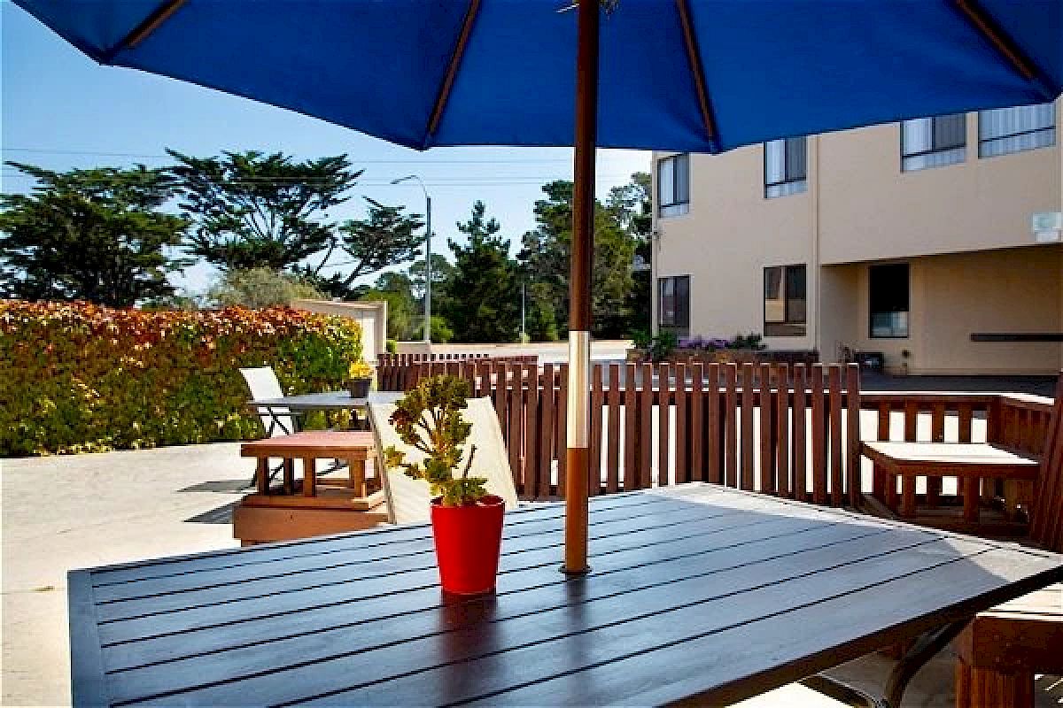 A patio with a wooden table, blue umbrella, and red plant pot. Background has chairs, a fence, and buildings, under a clear sky.