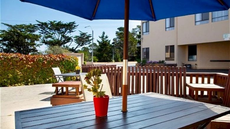 A patio with a wooden table, blue umbrella, and red plant pot. Background has chairs, a fence, and buildings, under a clear sky.