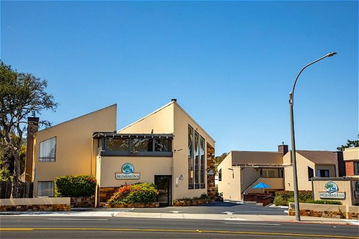 A modern building with large glass windows, beige walls, landscaping, and signs for "Munras Inn," located along a street with trees nearby.