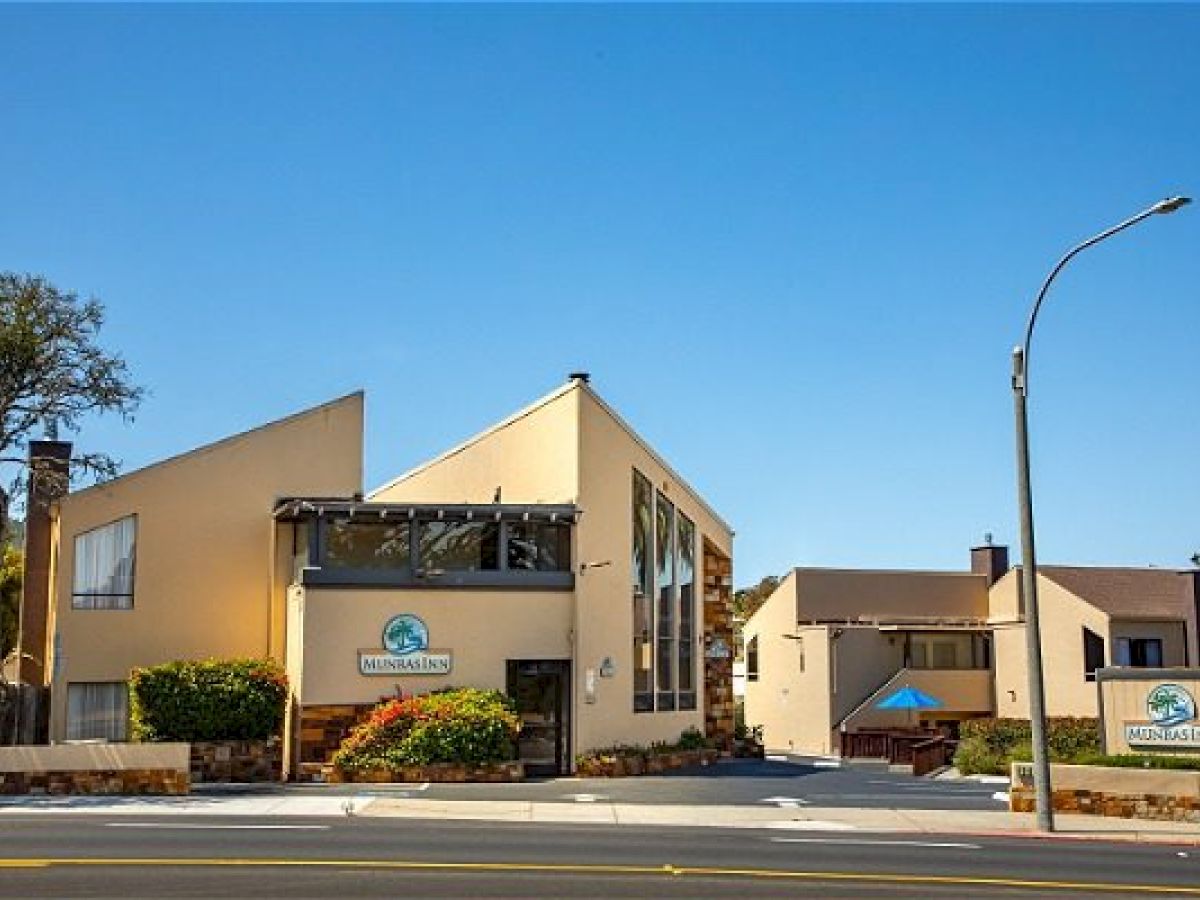 A modern building with large glass windows, beige walls, landscaping, and signs for "Munras Inn," located along a street with trees nearby.