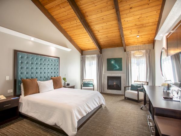 A cozy hotel room with a large bed, wooden ceiling, fireplace, and modern decor, featuring a desk and seating area by the window.