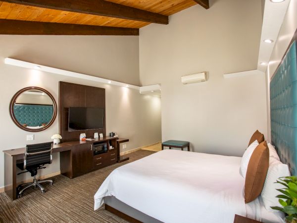 The image shows a modern hotel room with a bed, desk, chair, TV, and mirror. The decor is minimalist with a blue headboard and wooden ceiling.