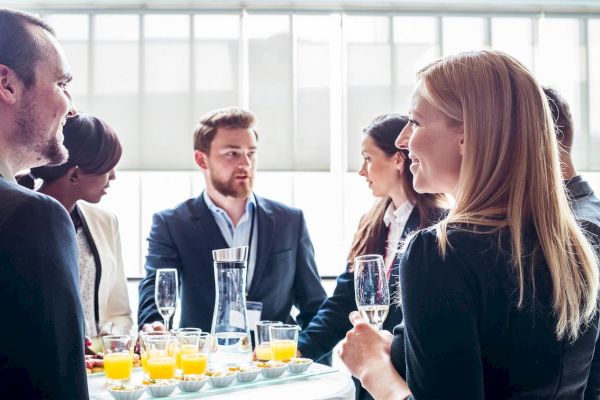 A group of people in formal attire are socializing around a table with drinks and snacks in a bright room.