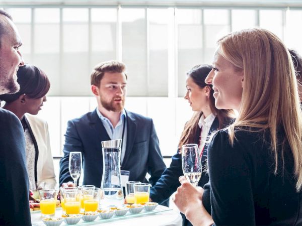 A group of people in formal attire are socializing around a table with drinks and snacks in a bright room.